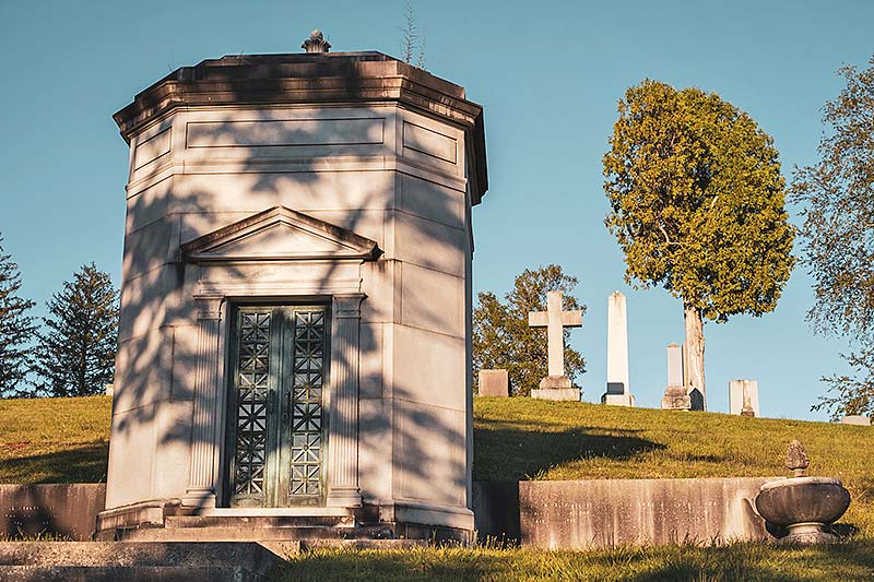 Mausoleum with headstones, trees, and blue sky in background.